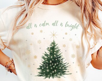 May include: A white t-shirt with the text "all is calm, all is bright" and a green watercolor Christmas tree with gold stars and snowflakes.