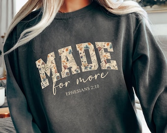 Könnte beinhalten: Dunkelgraues Sweatshirt mit dem Text "MADE for more" in floralem Design. Der Text "Epheser 2:10" steht darunter. Das Sweatshirt hat eine lockere Passform.