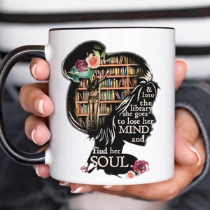 Könnte beinhalten: Eine weiße Keramiktasse mit schwarzem Henkel. Die Tasse zeigt eine Schwarzweißillustration einer Frau, die in einer Bibliothek mit Bücherregalen voller Bücher steht. Der Text lautet "& Into the library she goes to lose her MIND and find her SOUL."