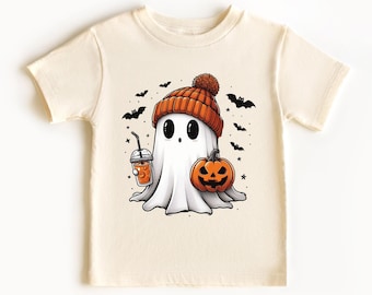 May include: A cream colored t-shirt with a cartoon ghost wearing an orange and white hat, holding a pumpkin and a cup of orange liquid. The ghost has big eyes and a small mouth. There are bats and stars around the ghost.