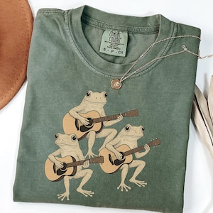Może przedstawiać: Zielona koszulka w kolorze mchu z grafiką przedstawiającą trzy żaby grające na gitarach. Koszulka ma okrągły dekolt i krótkie rękawy. Srebrny naszyjnik z wisiorkiem jest zarzucony na koszulkę. Koszulka jest wykonana z miękkiego, wygodnego materiału.