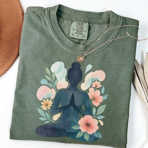 以下が含まれることがあります： モスグリーンのComfort Colors Tシャツ。水彩画のデザインで、ヨガのポーズをとる人物が花や抽象的な形に囲まれています。小さなペンダントが付いたシルバーのネックレスがシャツの上にあります。シャツのサイズはS-P-CHです。