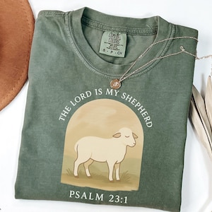 Op de afbeelding: Een groen T-shirt met een witte afbeelding van een schaap en de tekst "THE LORD IS MY SHEPHERD" en "PSALM 23:1".