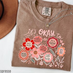 May include: A brown t-shirt with a pink and red floral design and the text "it's OKAY to" with phrases like "make mistakes", "start over", "have strong emotions", "be different", "ask for help", and "have bad days" inside the flowers.