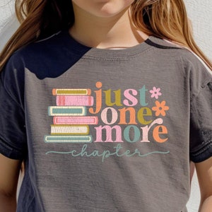 May include: A dark gray t-shirt with a colorful graphic of stacked books and the text "just one more chapter". The shirt is being worn by a child with pink round sunglasses.