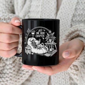 Black coffee mug with skeleton reading while lounging in a boho style room with the words Just One More Chapter
