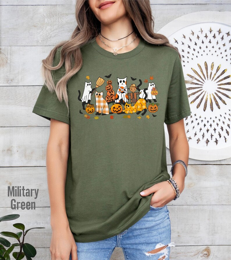 May include: Olive green t-shirt featuring a Halloween-themed graphic. The design includes cartoon cats dressed as ghosts, pumpkins, and scarecrows. The text "Military Green" is printed on the left side of the shirt.