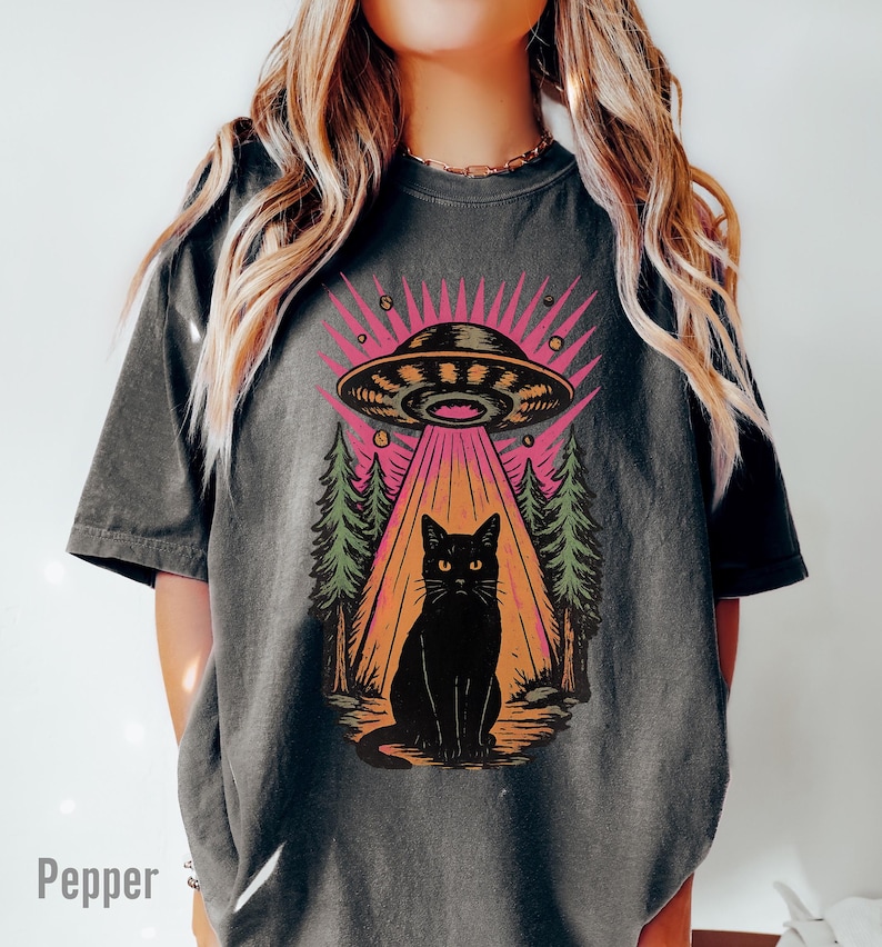 May include: Dark grey t-shirt featuring a black cat under a UFO with a pink and orange beam. The design includes green trees and the word "Pepper" at the bottom left. The shirt has short sleeves.