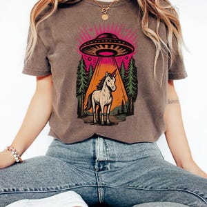 May include: A brown t-shirt featuring a graphic of a unicorn standing in a forest, illuminated by a UFO beam. The design includes a pink and orange color scheme with a forest backdrop.
