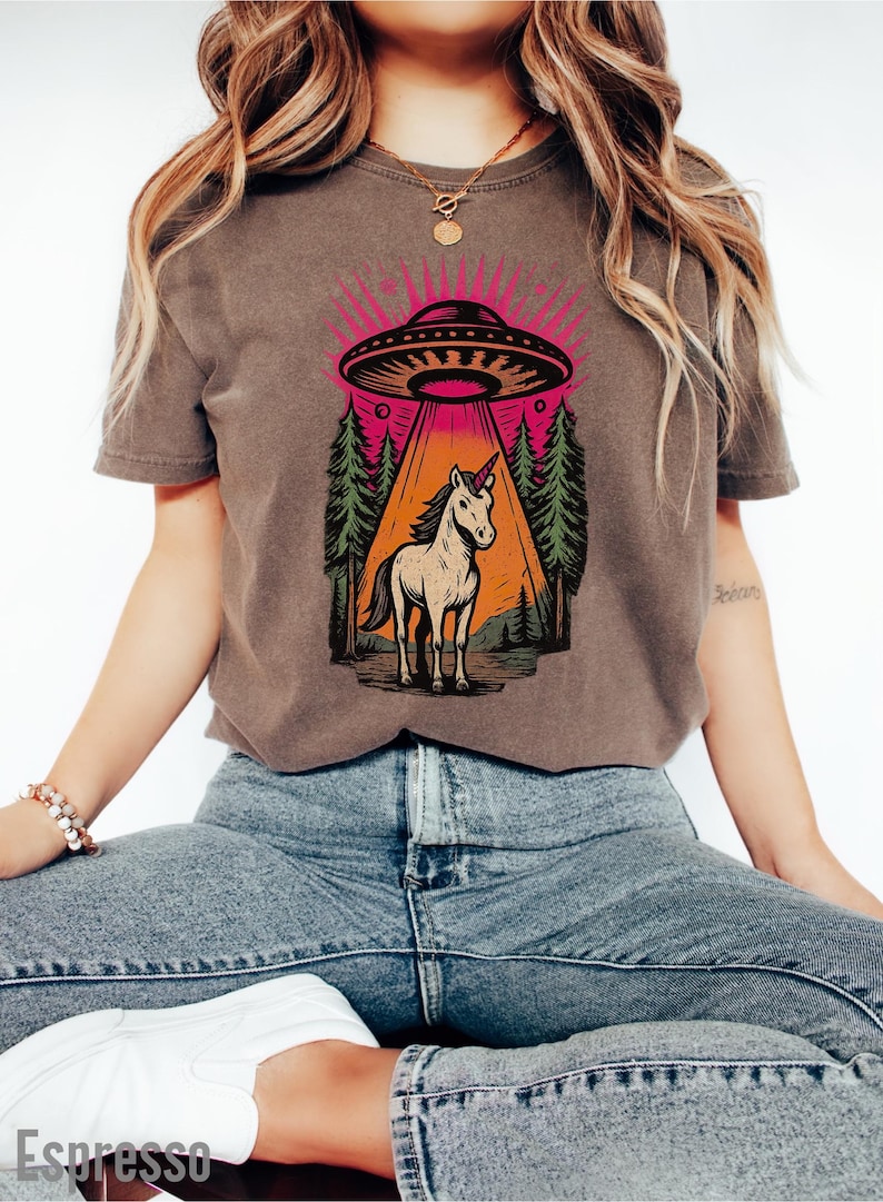 May include: A brown t-shirt featuring a graphic of a unicorn standing in a forest, illuminated by a UFO beam. The design includes a pink and orange color scheme with a forest backdrop.