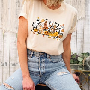 May include: Cream-colored t-shirt featuring a Halloween-themed graphic. The design includes cartoon cats dressed as ghosts, pumpkins, and a scarecrow, with bats and autumn leaves. The shirt is paired with light blue jeans.