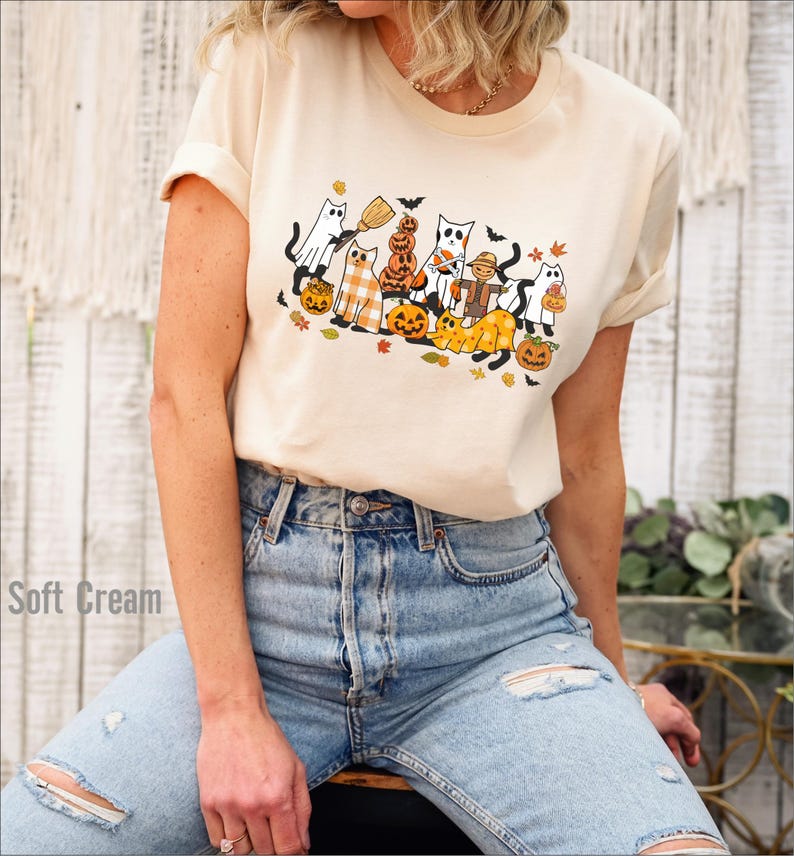 May include: Cream-colored t-shirt featuring a Halloween-themed graphic. The design includes cartoon cats dressed as ghosts, pumpkins, and a scarecrow, with bats and autumn leaves. The shirt is paired with light blue jeans.
