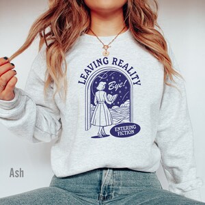 May include: A gray sweatshirt with a blue graphic of a woman reading a book in a doorway. The text on the graphic reads "Leaving Reality Bye! Entering Fiction".