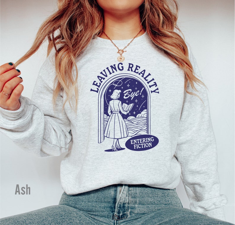 May include: A gray sweatshirt with a blue graphic of a woman reading a book in a doorway. The text on the graphic reads "Leaving Reality Bye! Entering Fiction".