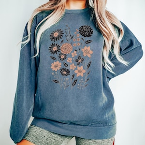 May include: Blue jean-colored sweatshirt featuring a floral design in black and peach tones. The crew neck sweatshirt has long sleeves and a relaxed fit. The text "Blue Jean" is visible at the bottom right.