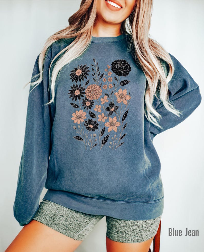 May include: Blue jean-colored sweatshirt featuring a floral design in black and peach tones. The crew neck sweatshirt has long sleeves and a relaxed fit. The text "Blue Jean" is visible at the bottom right.