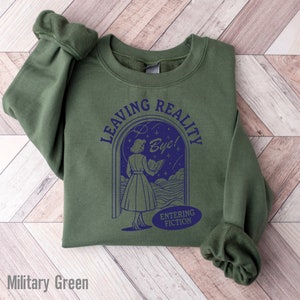 May include: A military green sweatshirt with a graphic of a woman walking through a doorway with the text "Leaving Reality Bye! Entering Fiction".