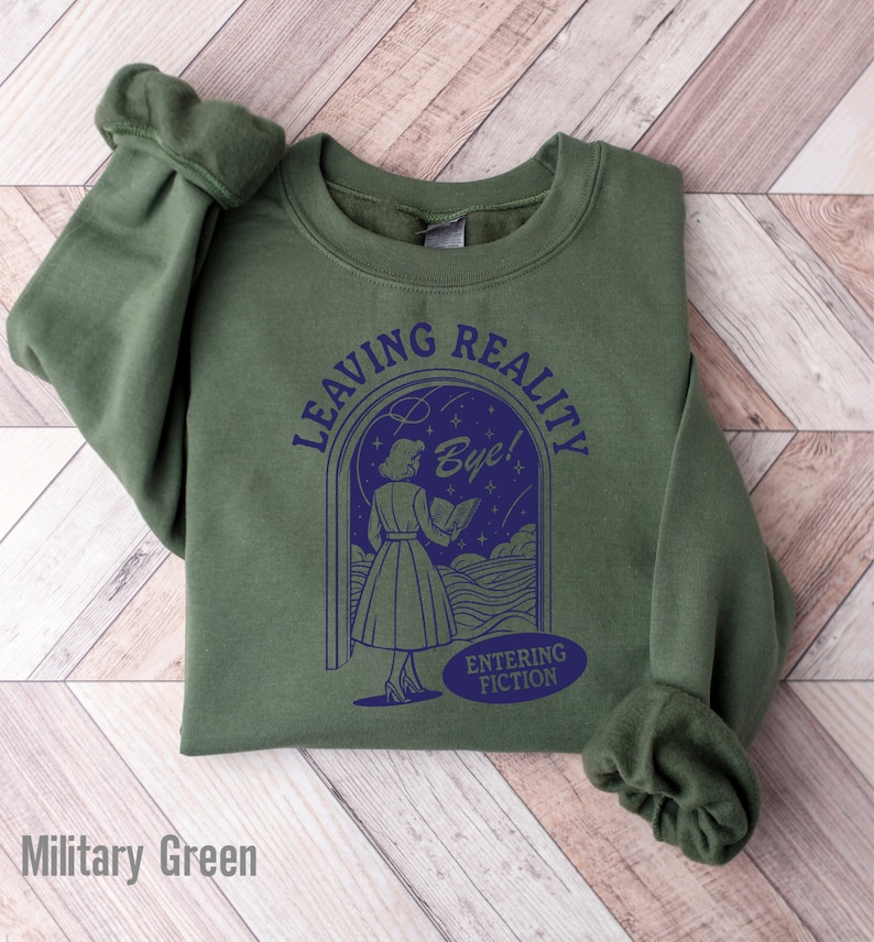 May include: A military green sweatshirt with a graphic of a woman walking through a doorway with the text "Leaving Reality Bye! Entering Fiction".