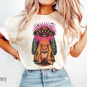 May include: Ivory-colored t-shirt featuring a colorful graphic of a fox sitting beneath a UFO. The UFO is emitting a beam of light, and the design includes trees and planets. The word "Ivory" is printed at the bottom left.