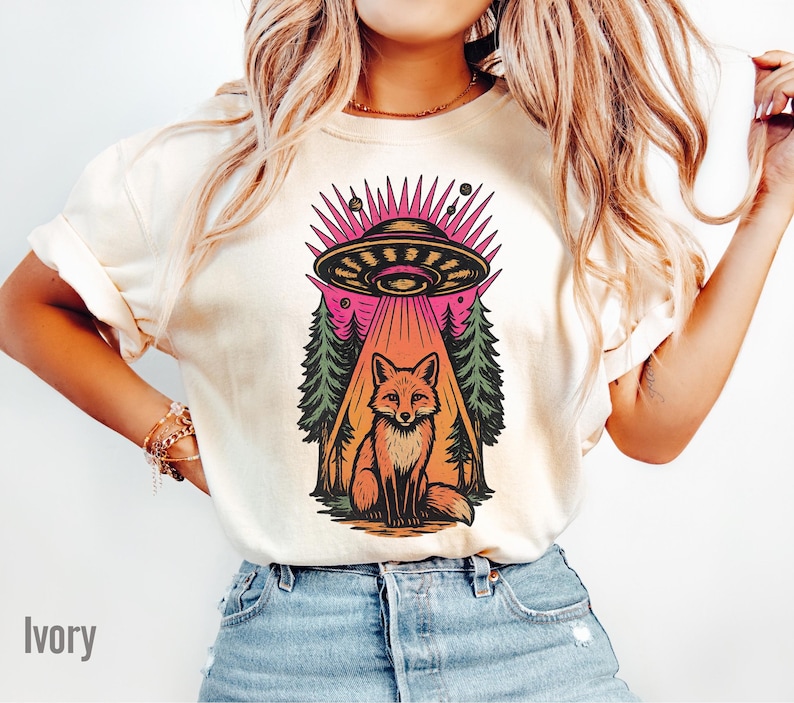 May include: Ivory-colored t-shirt featuring a colorful graphic of a fox sitting beneath a UFO. The UFO is emitting a beam of light, and the design includes trees and planets. The word "Ivory" is printed at the bottom left.