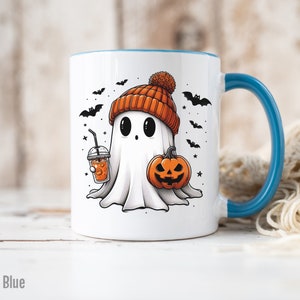 May include: White ceramic mug with a light blue rim and handle. The mug features a cartoon ghost wearing an orange knit hat and holding a pumpkin and a cup of orange liquid. The background includes black bats and stars.