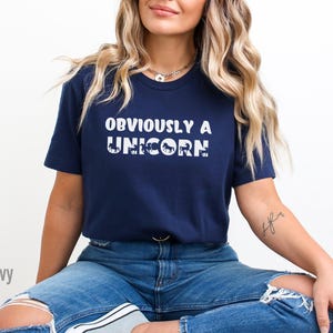 May include: A navy blue t-shirt with the text "OBVIOUSLY A UNICORN" in white. The word "UNICORN" is made up of a series of small white unicorn silhouettes.