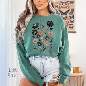May include: Light green sweatshirt featuring a floral design in black and tan. The sweatshirt has a crew neck and long sleeves. The words "Light Green" are printed on the bottom left of the image. The sweatshirt is paired with denim shorts.