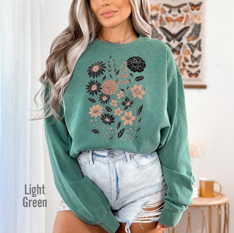 May include: Light green sweatshirt featuring a floral design in black and tan. The sweatshirt has a crew neck and long sleeves. The words "Light Green" are printed on the bottom left of the image. The sweatshirt is paired with denim shorts.