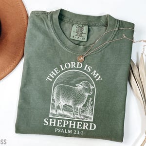 Puede incluir: Una camiseta verde con un gr&aacute;fico blanco de una oveja y el texto "The Lord is my Shepherd Psalm 23:1".
