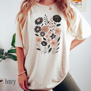 May include: Ivory-colored t-shirt featuring a floral design with black and peach-colored flowers and green leaves. The shirt has a relaxed fit and short sleeves. The word "Ivory" is printed on the bottom left.