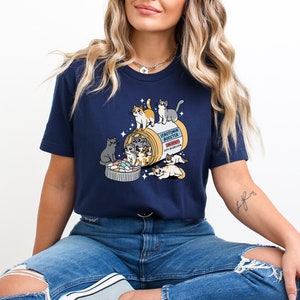 May include: A navy blue t-shirt with a graphic of a pill bottle labeled "Serotonin Booster" with several cartoon cats spilling out of it. The text "meowdication" is printed on the pill bottle.