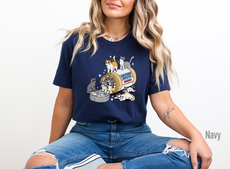May include: A navy blue t-shirt with a graphic of a pill bottle labeled "Serotonin Booster" with several cartoon cats spilling out of it. The text "meowdication" is printed on the pill bottle.