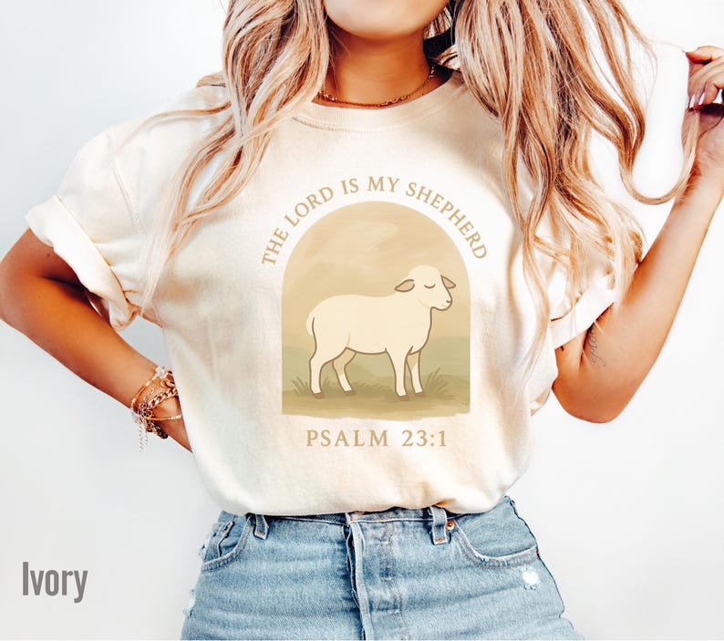 Op de afbeelding: Een ivoorwit T-shirt met een afbeelding van een schaap in een weiland met de tekst "The Lord is my Shepherd, Psalm 23:1".