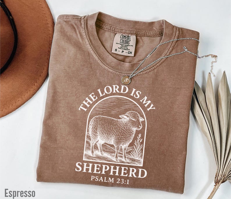 Puede incluir: Una camiseta marr&oacute;n con un dise&ntilde;o gr&aacute;fico blanco que dice "The Lord is my Shepherd" y "Psalm 23:1". El gr&aacute;fico presenta una oveja estilizada en un campo.