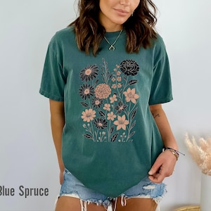 Peut inclure: Un t-shirt de couleur sarcelle avec un motif floral noir et p&ecirc;che. Le graphisme comprend divers types de fleurs et de feuillage. Le texte "Blue Spruce" est imprim&eacute; en bas de l'image. Le t-shirt est en mati&egrave;re souple.
