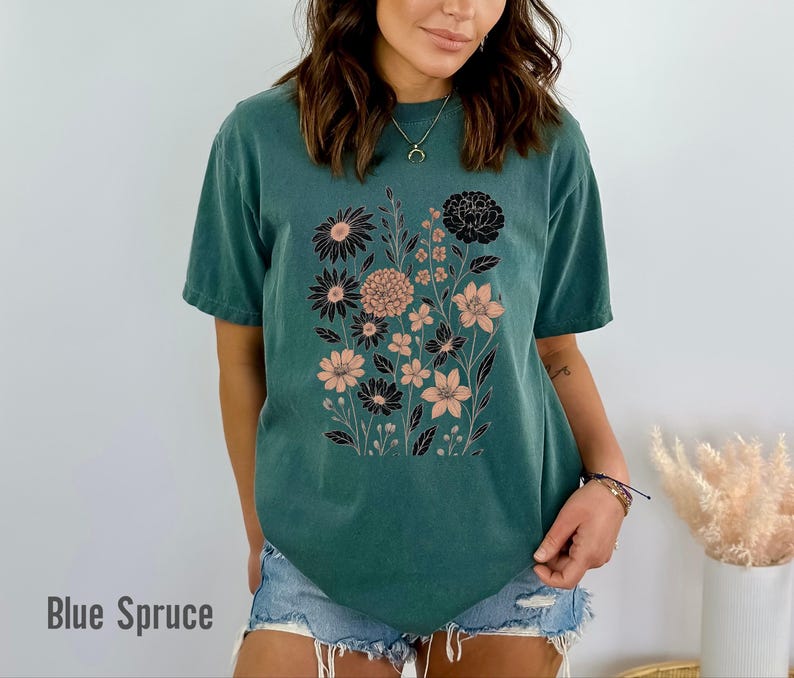 Peut inclure: Un t-shirt de couleur sarcelle avec un motif floral noir et p&ecirc;che. Le graphisme comprend divers types de fleurs et de feuillage. Le texte "Blue Spruce" est imprim&eacute; en bas de l'image. Le t-shirt est en mati&egrave;re souple.