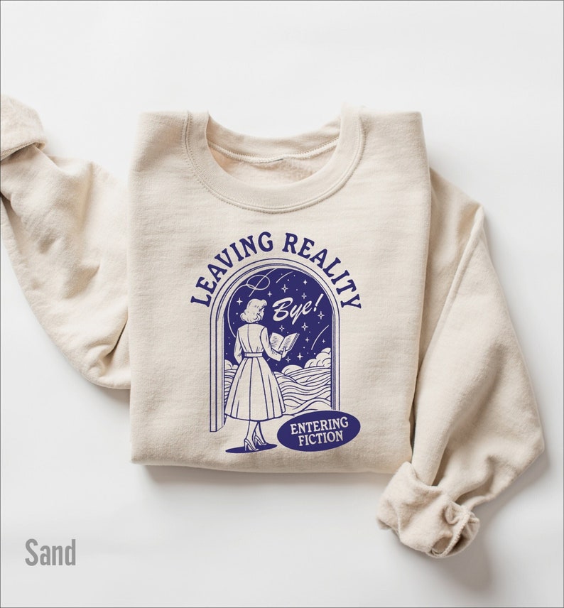 May include: A sand-colored sweatshirt with a blue graphic of a woman walking through a doorway with the text "Leaving Reality Bye! Entering Fiction".