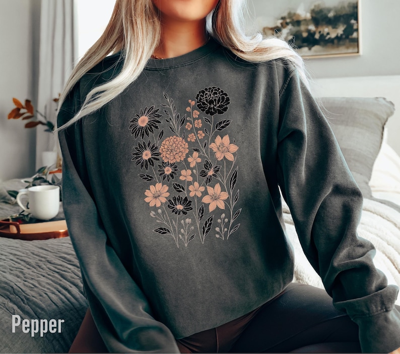May include: A dark gray sweatshirt with a floral design. The design features black and peach-colored flowers with green stems and leaves. The word "Pepper" is printed at the bottom left corner of the image.