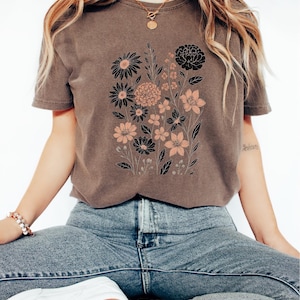 Peut inclure: T-shirt marron avec un motif floral noir et p&ecirc;che. Le graphisme comprend divers types de fleurs et de feuillage. Le t-shirt est port&eacute; avec un jean bleu et des baskets blanches. Le mot "Espresso" est imprim&eacute; en bas.