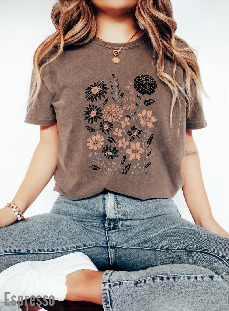 Peut inclure: T-shirt marron avec un motif floral noir et p&ecirc;che. Le graphisme comprend divers types de fleurs et de feuillage. Le t-shirt est port&eacute; avec un jean bleu et des baskets blanches. Le mot "Espresso" est imprim&eacute; en bas.