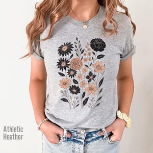 Op de afbeelding: Grijs Athletic Heather T-shirt met een bloemenontwerp in zwart en perzik. De afbeelding toont verschillende soorten bloemen en bladeren. Het shirt heeft korte mouwen en een ronde hals.
