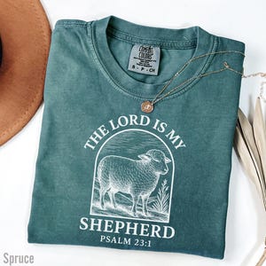 Puede incluir: Una camiseta azul turquesa con un gr&aacute;fico blanco de una oveja y el texto "The Lord is my Shepherd Psalm 23:1".