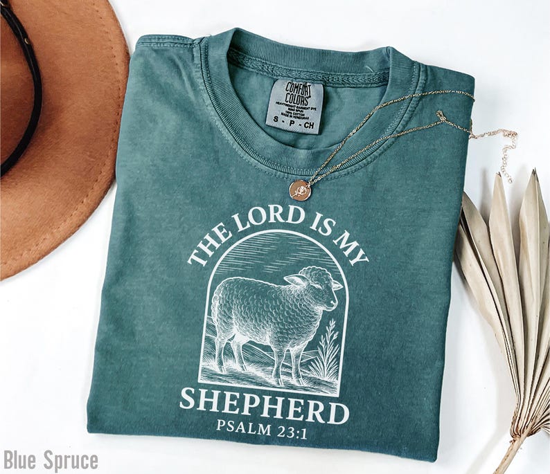 Puede incluir: Una camiseta azul turquesa con un gr&aacute;fico blanco de una oveja y el texto "The Lord is my Shepherd Psalm 23:1".