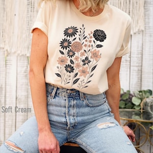 Op de afbeelding: Zacht cr&egrave;mekleurig T-shirt met een bloemenontwerp van zwarte en blush-kleurige bloemen en groene stelen. Het shirt wordt gecombineerd met lichtblauwe ripped jeans. De tekst "Soft Cream" is zichtbaar aan de linkerkant van de afbeelding.