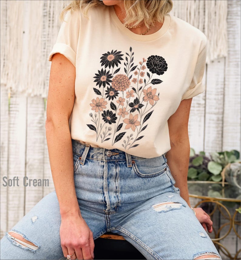 Op de afbeelding: Zacht cr&egrave;mekleurig T-shirt met een bloemenontwerp van zwarte en blush-kleurige bloemen en groene stelen. Het shirt wordt gecombineerd met lichtblauwe ripped jeans. De tekst "Soft Cream" is zichtbaar aan de linkerkant van de afbeelding.