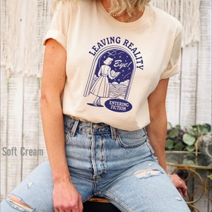 May include: A cream colored t-shirt with a blue graphic of a woman walking through a doorway with the text "Leaving Reality" and "Entering Fiction" and "Bye!"