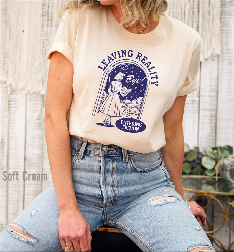 May include: A cream colored t-shirt with a blue graphic of a woman walking through a doorway with the text "Leaving Reality" and "Entering Fiction" and "Bye!"