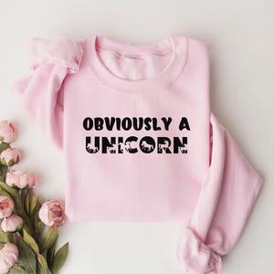 May include: A light pink sweatshirt with the text "OBVIOUSLY A UNICORN" in black lettering. The text is surrounded by small black unicorn silhouettes.