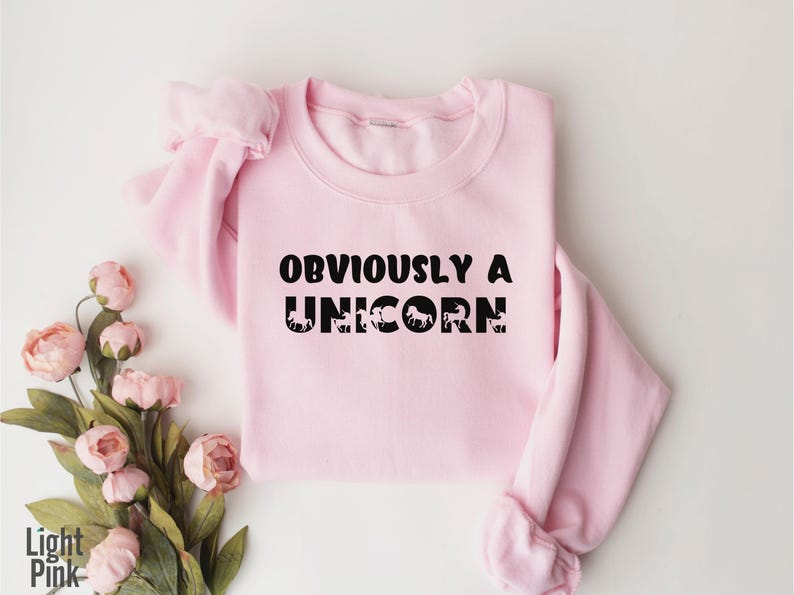 May include: A light pink sweatshirt with the text "OBVIOUSLY A UNICORN" in black lettering. The text is surrounded by small black unicorn silhouettes.