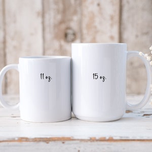 Op de afbeelding: Twee witte keramische mokken met zwarte letters. De kleinere mok is 11 oz en de grotere mok is 15 oz.